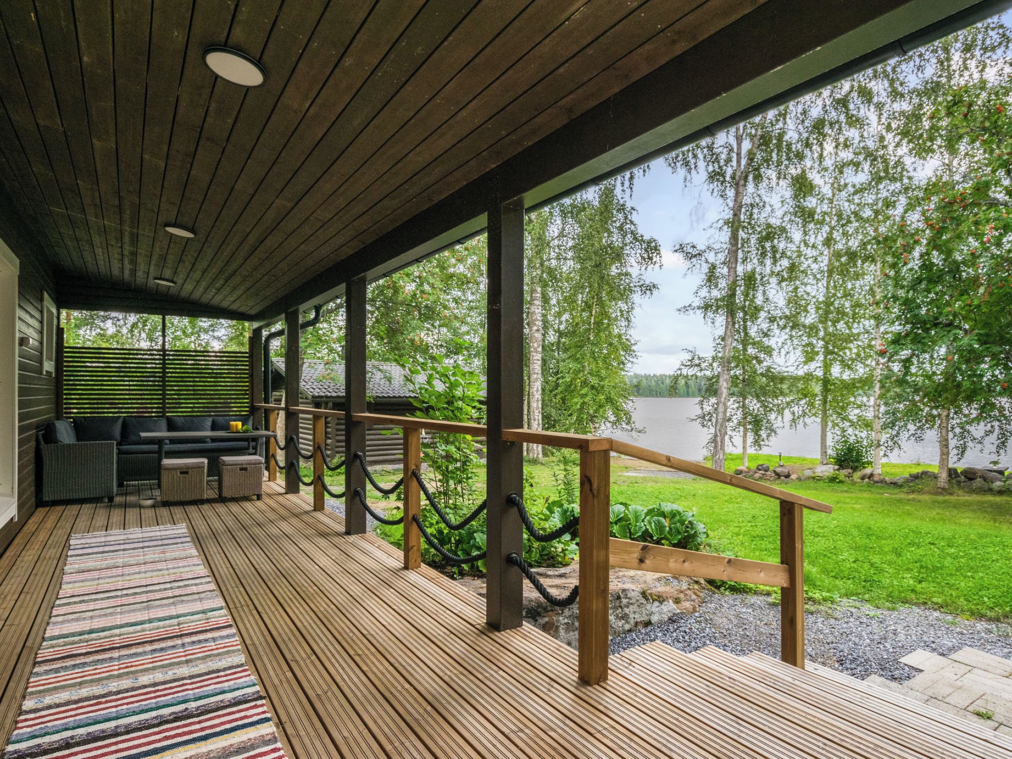 Une terrasse extérieure couverte, dotée d’un plancher en bois, d’un tapis à rayures et d’un espace salon avec un canapé et des fauteuils.