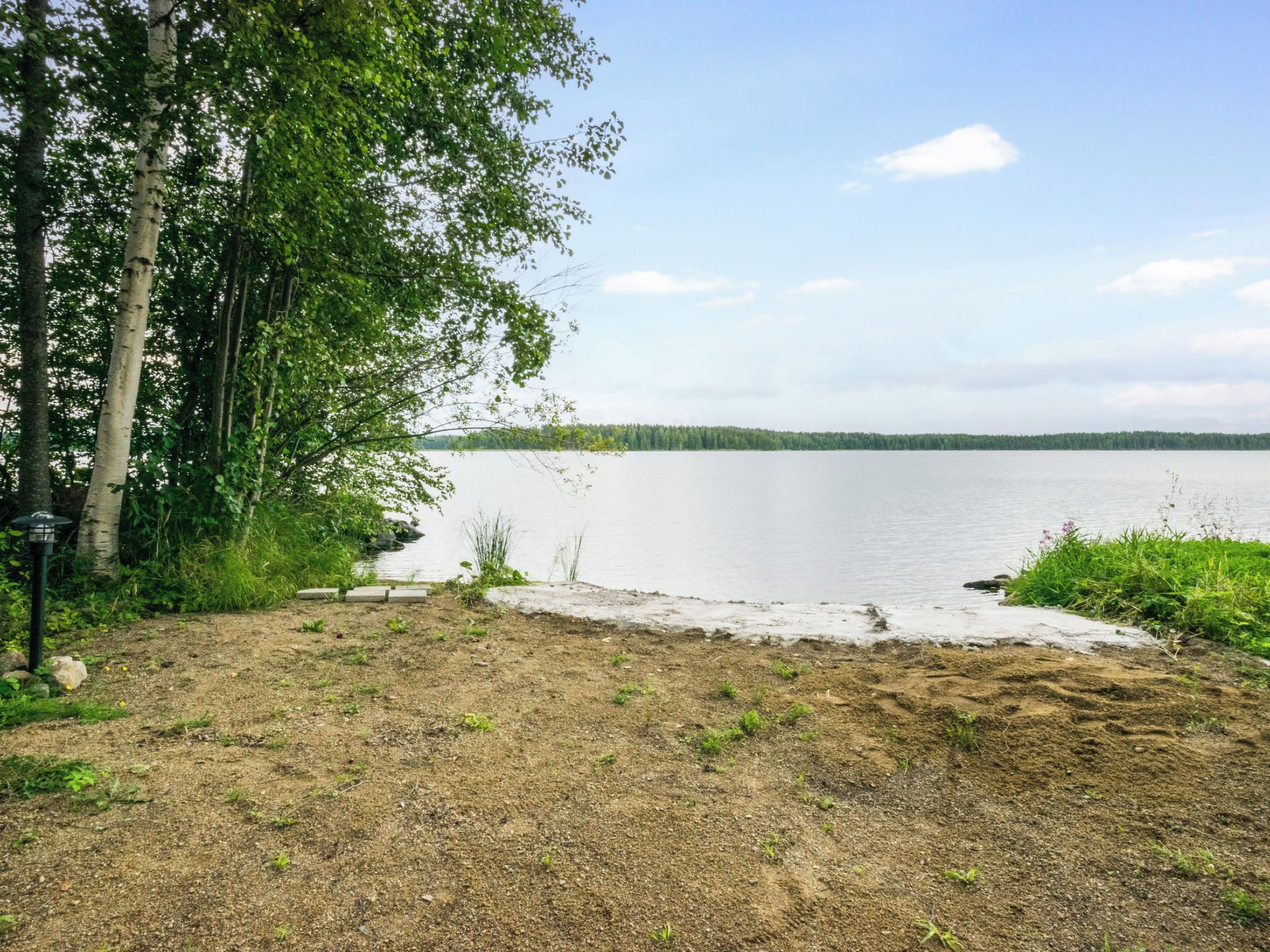 Vista de un lago con un sendero de tierra que lleva a una zona arenosa, rodeada de árboles y un cielo despejado.