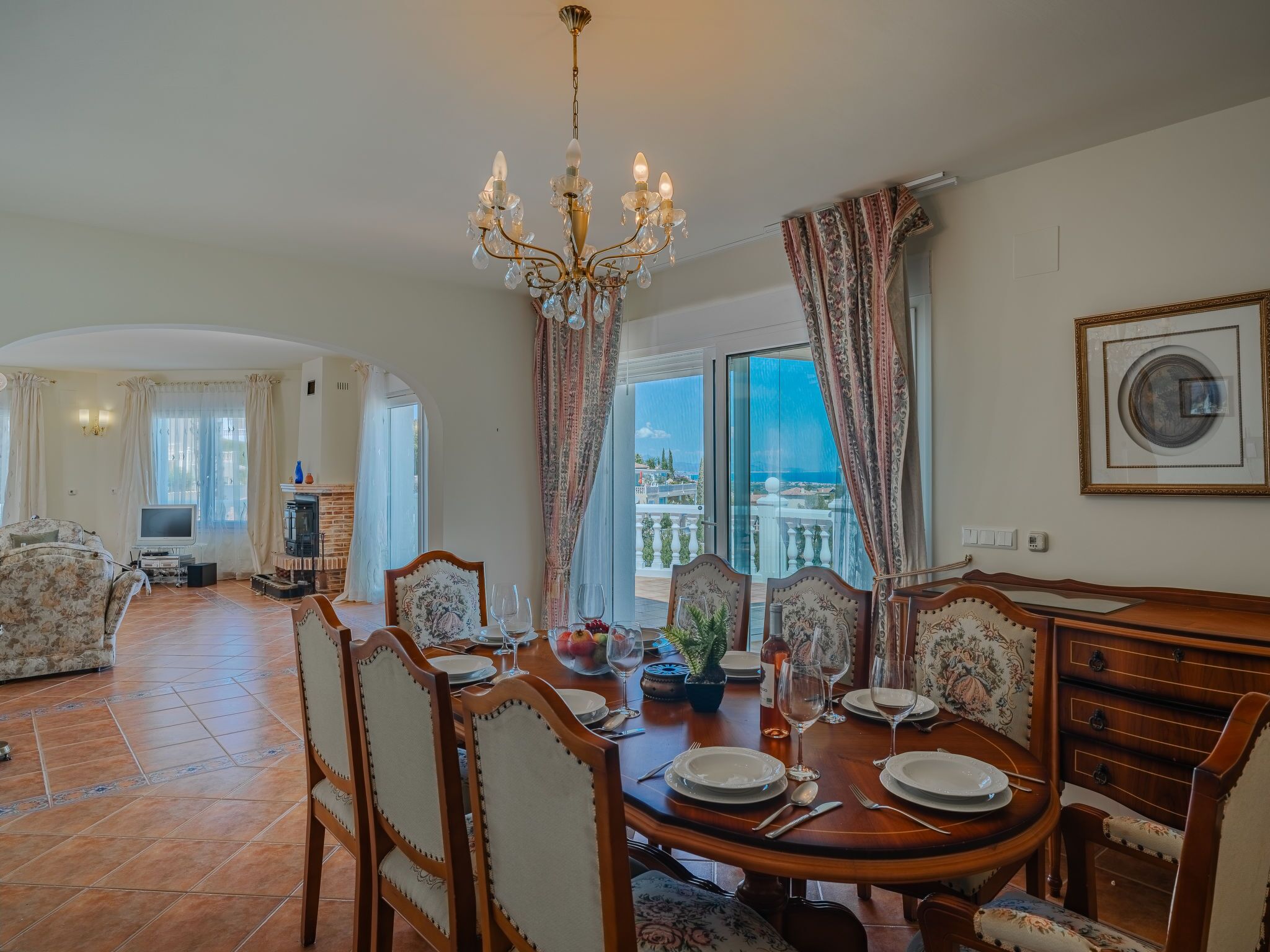 Un coin repas avec une table ronde dressée pour quatre personnes, entourée de chaises à motifs floraux. Un lustre est suspendu au-dessus, et une grande fenêtre offre une vue sur la mer.