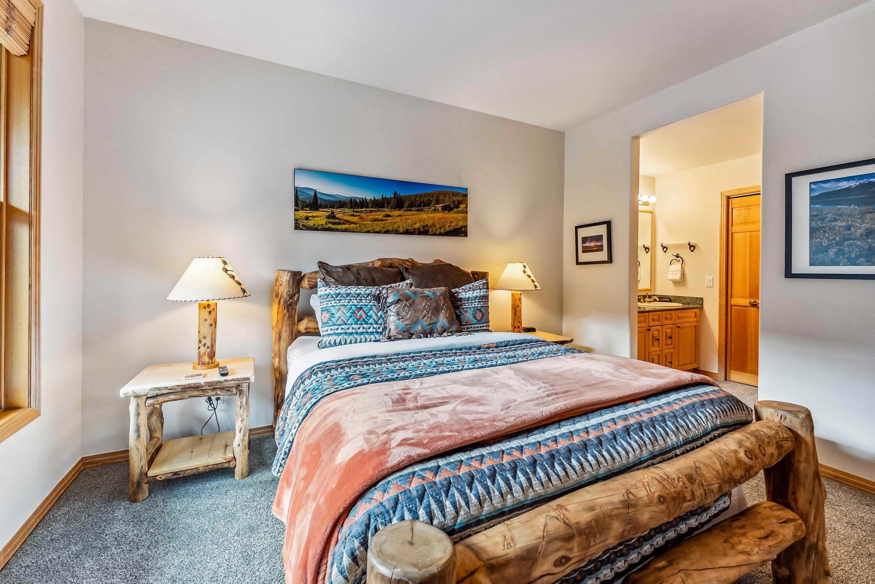 A bedroom with a bed, two bedside tables, a nightstand, a wooden bench, a wall-mounted picture, and a door leading to another room.