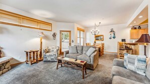 A spacious living room with a grey sofa, wooden coffee table, and a patterned armchair. A dining area with wooden chairs and a chandelier is visible in the background. - Breckenridge Condo with Hot Tub Access (Breckenridge)