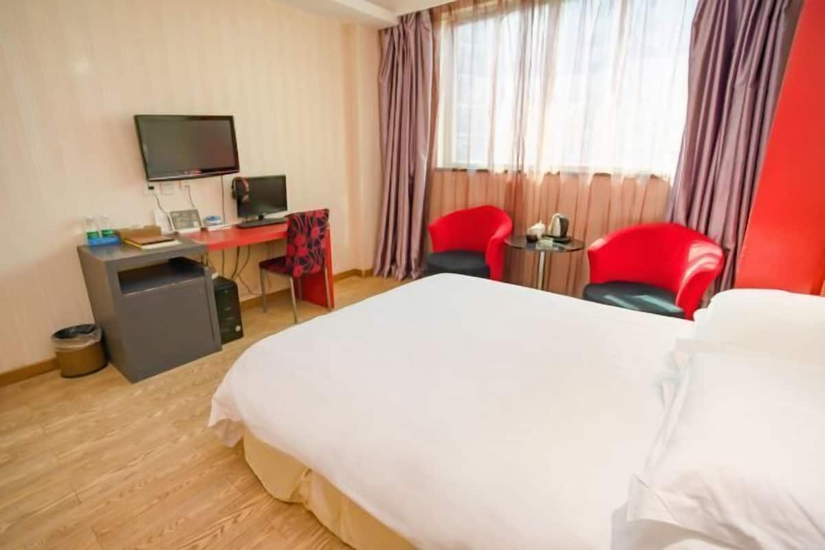 A hotel room with a bed, a desk with a computer, two red chairs, and a television.