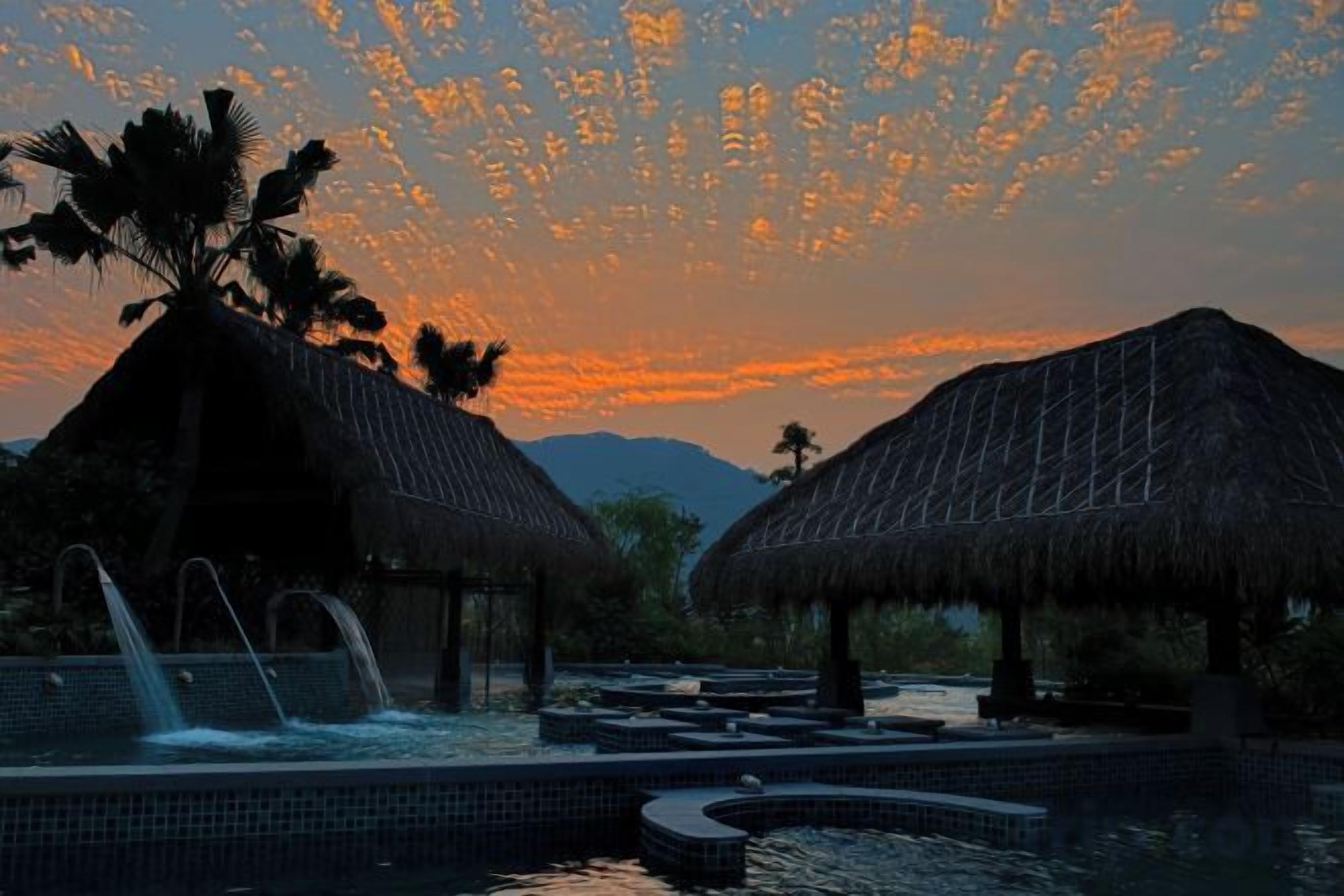 A resort with thatched-roof structures, a water feature, and a sunset view.