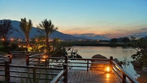Una terraza de madera con barandillas que da a una masa de agua tranquila, rodeada de follaje y montañas al fondo.