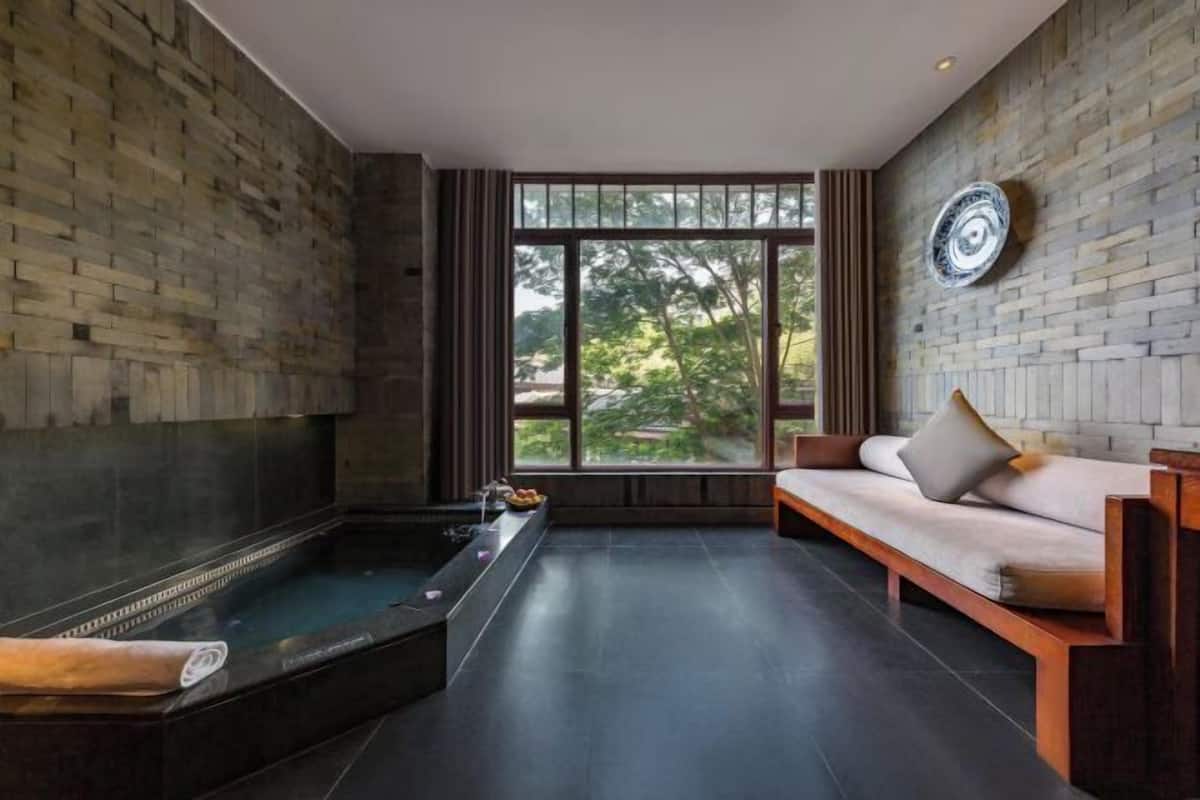 A spa room with a stone wall, a wooden bench, a massage table, and a window with a view of greenery.