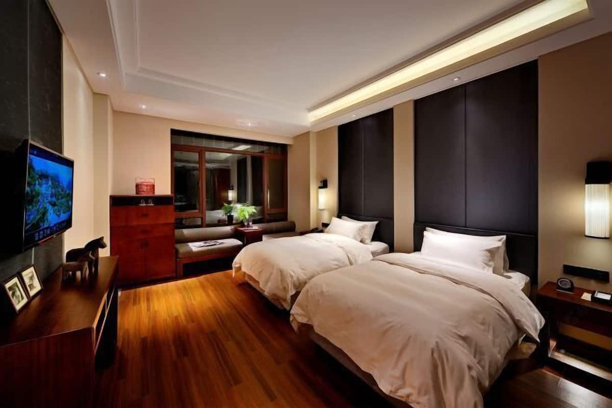 A modern hotel room with a wooden floor, two beds, a TV, a desk, and a large window.