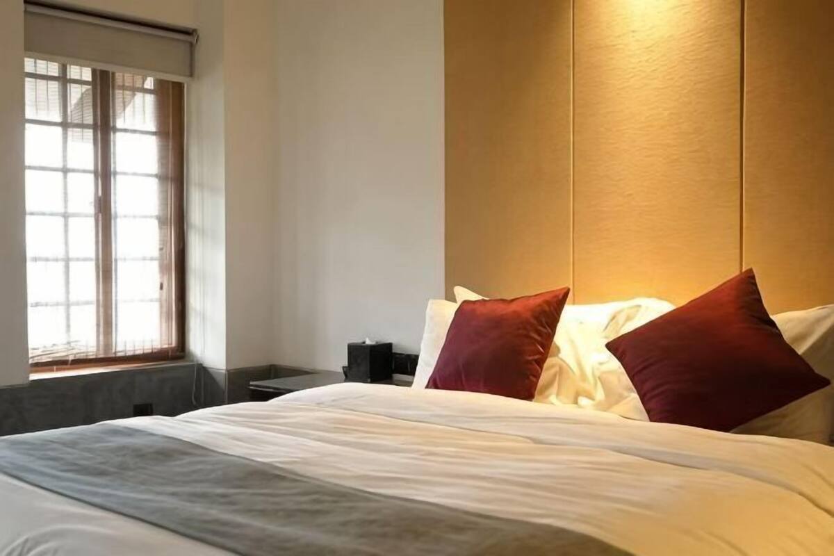 A hotel room with a bed, pillows, a nightstand, a window with blinds, and a wall-mounted light.