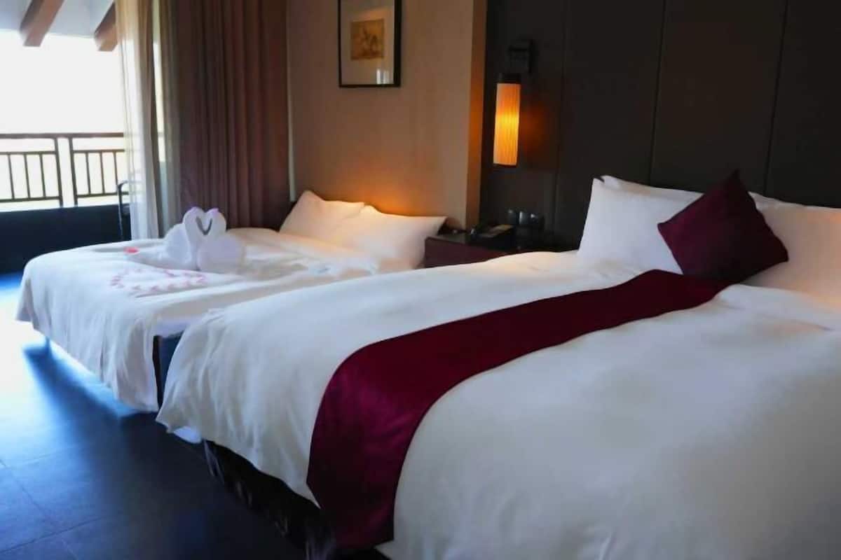 A hotel room with two beds, a red throw blanket, white pillows, a nightstand, a lamp, and a framed picture on the wall.