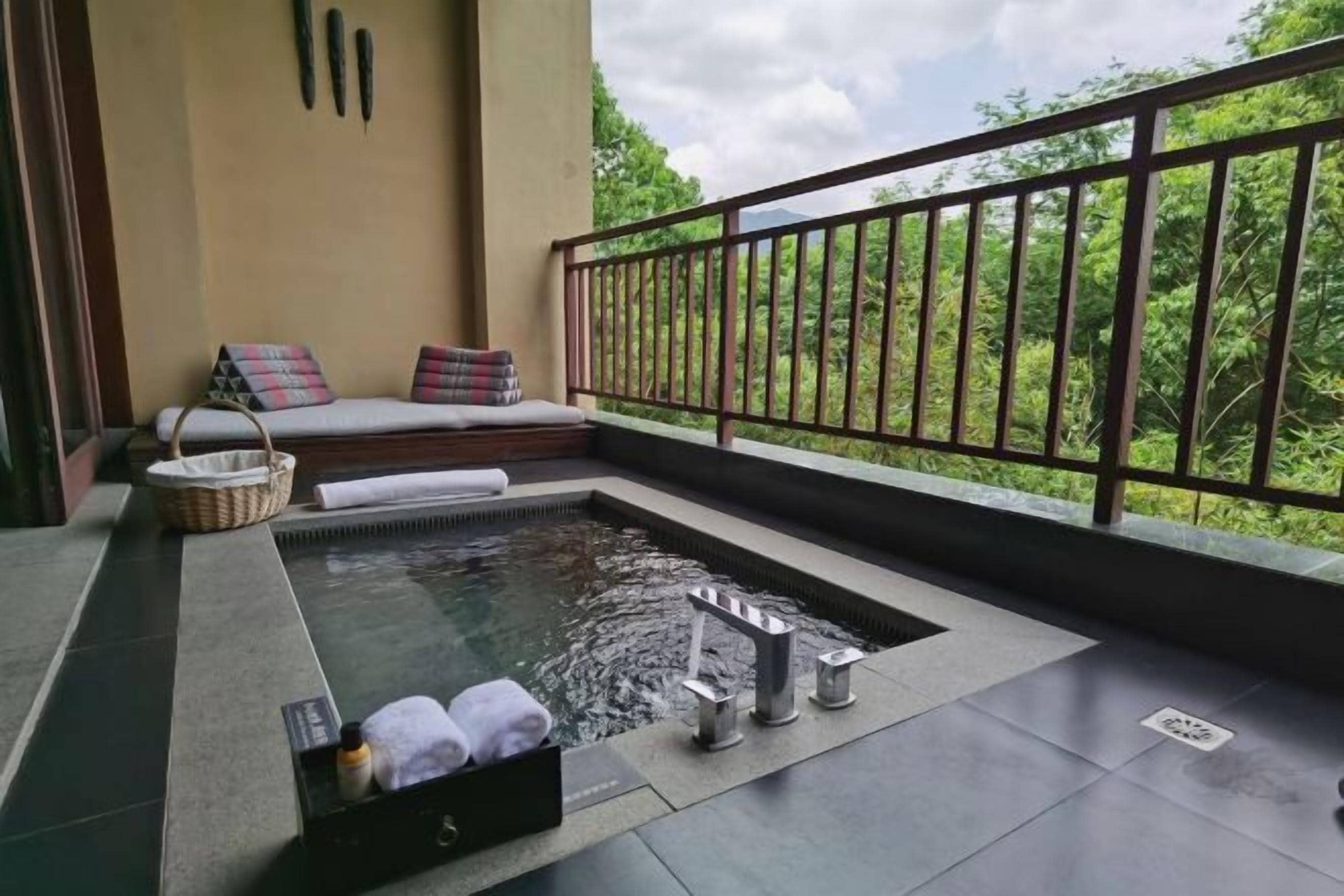Un balcón con un jacuzzi, una zona de estar con cojines y vistas a la vegetación.