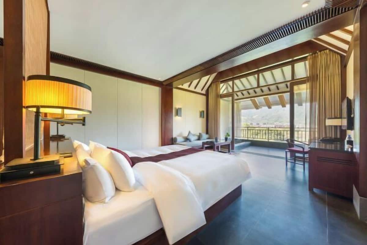 A hotel room with a large bed, a desk, a chair, a lamp, and a balcony with a view.