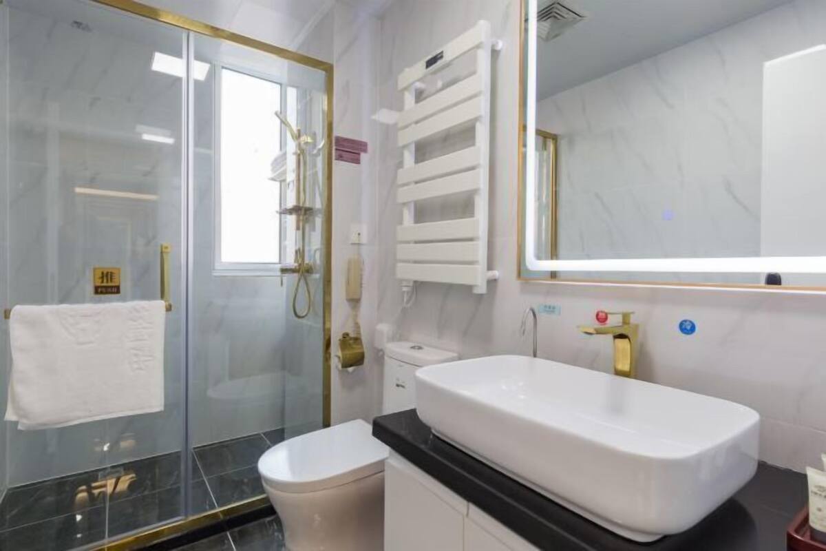A modern bathroom with a glass shower enclosure, a white toilet, a large sink with a gold faucet, and a mirror with a gold frame.