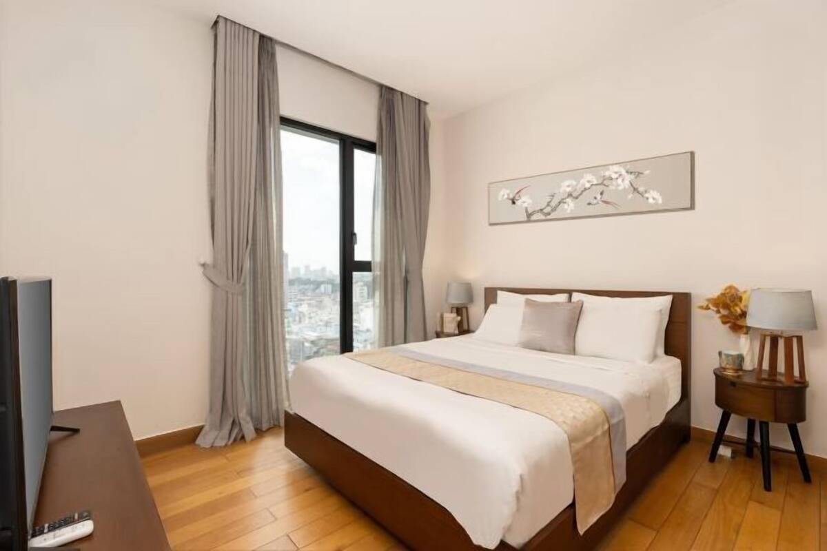 Ein Hotelzimmer mit einem großen Bett, einem Fernseher, einem Nachttisch, einer Lampe und einem Fenster mit Vorhängen.