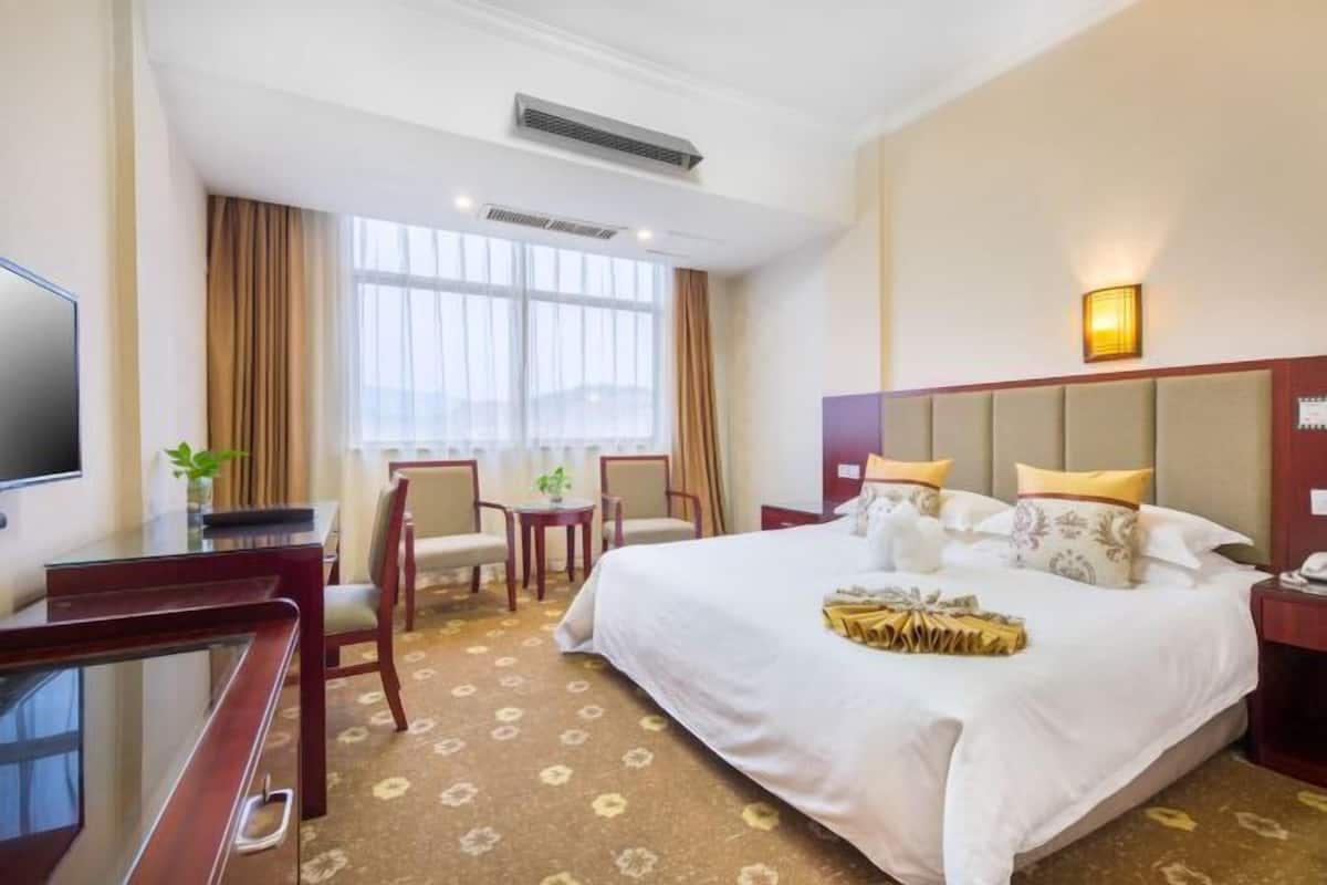 A hotel room with a large bed, a desk, chairs, a TV, and a window with curtains.