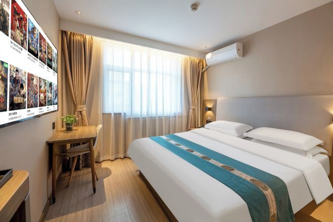 A hotel room with a bed, a desk, a chair, a window with curtains, and wall-mounted air conditioning.