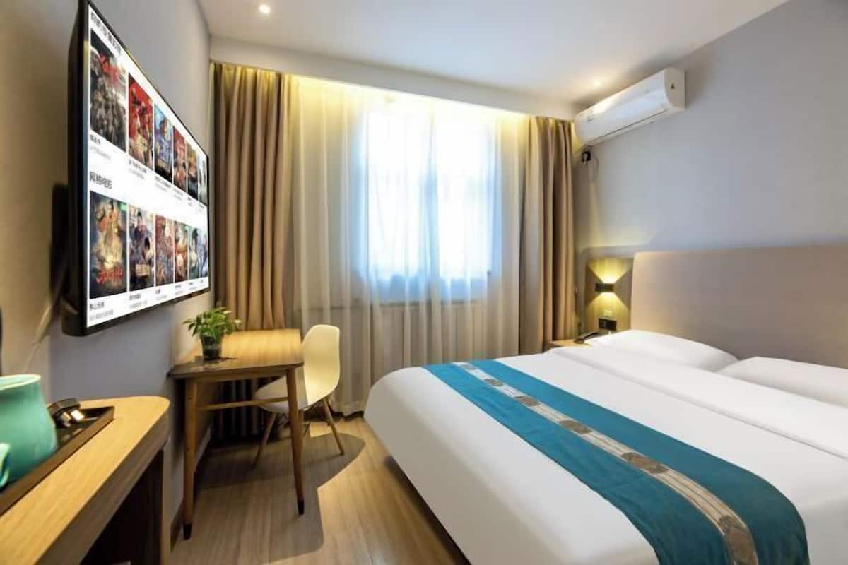 A hotel room with a bed, a desk, a chair, a TV mounted on the wall, and a window with curtains.