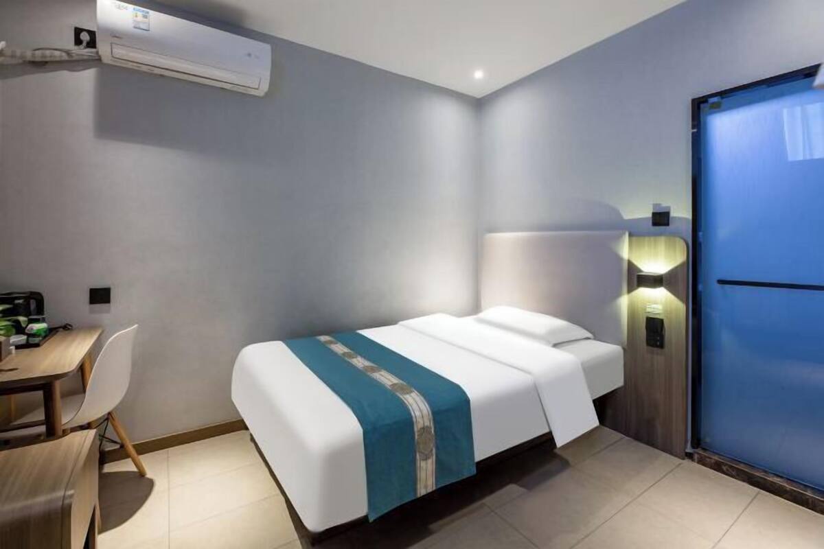 A hotel room with a bed, a desk, a chair, an air conditioning unit, and a door.