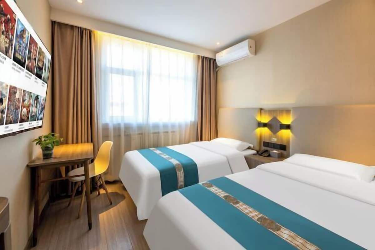 A hotel room with two beds, a desk, a chair, a wall-mounted air conditioner, and a wall-mounted lamp.