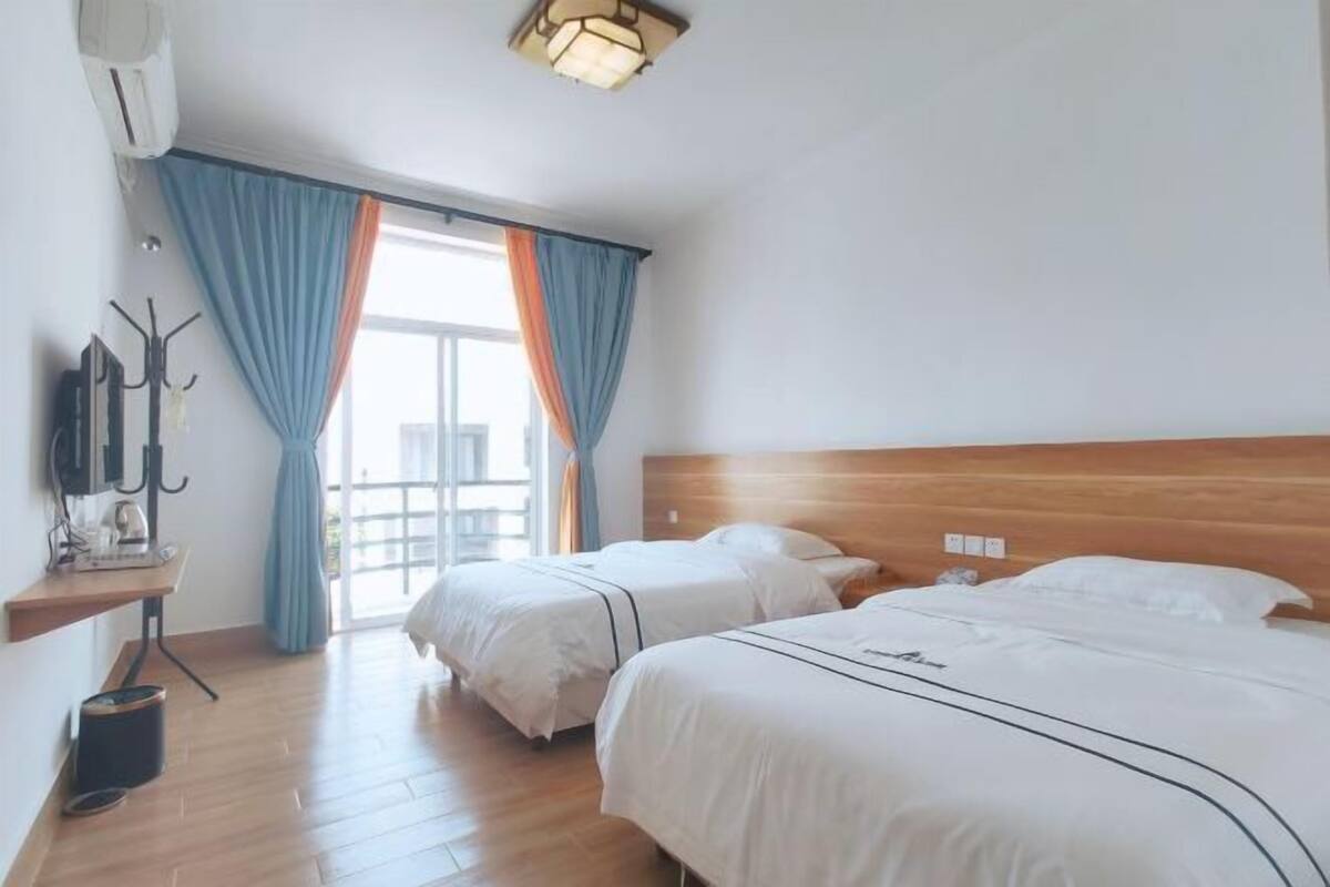 Ein Hotelzimmer mit zwei Betten, einem Holzkopfteil, einem Schreibtisch mit Stuhl und einem Fenster mit blauen Vorhängen.