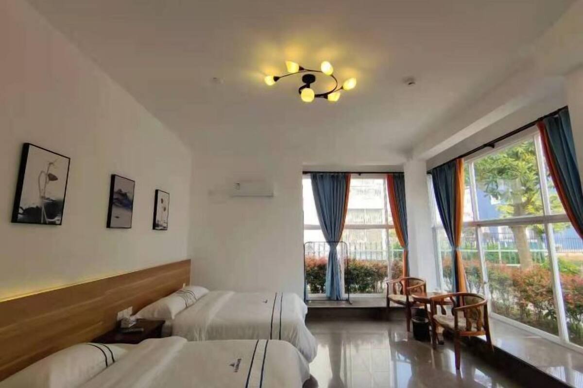 Ein Hotelzimmer mit zwei Betten, einem Holzkopfstück, einer Deckenleuchte, einem Fenster mit Vorhängen und Blick auf Grünfläche.