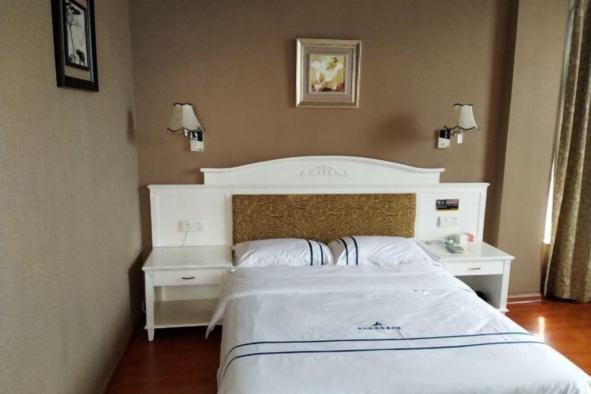 A hotel room with a white bed, a headboard with a textured design, two bedside tables with lamps, and a framed picture on the wall.