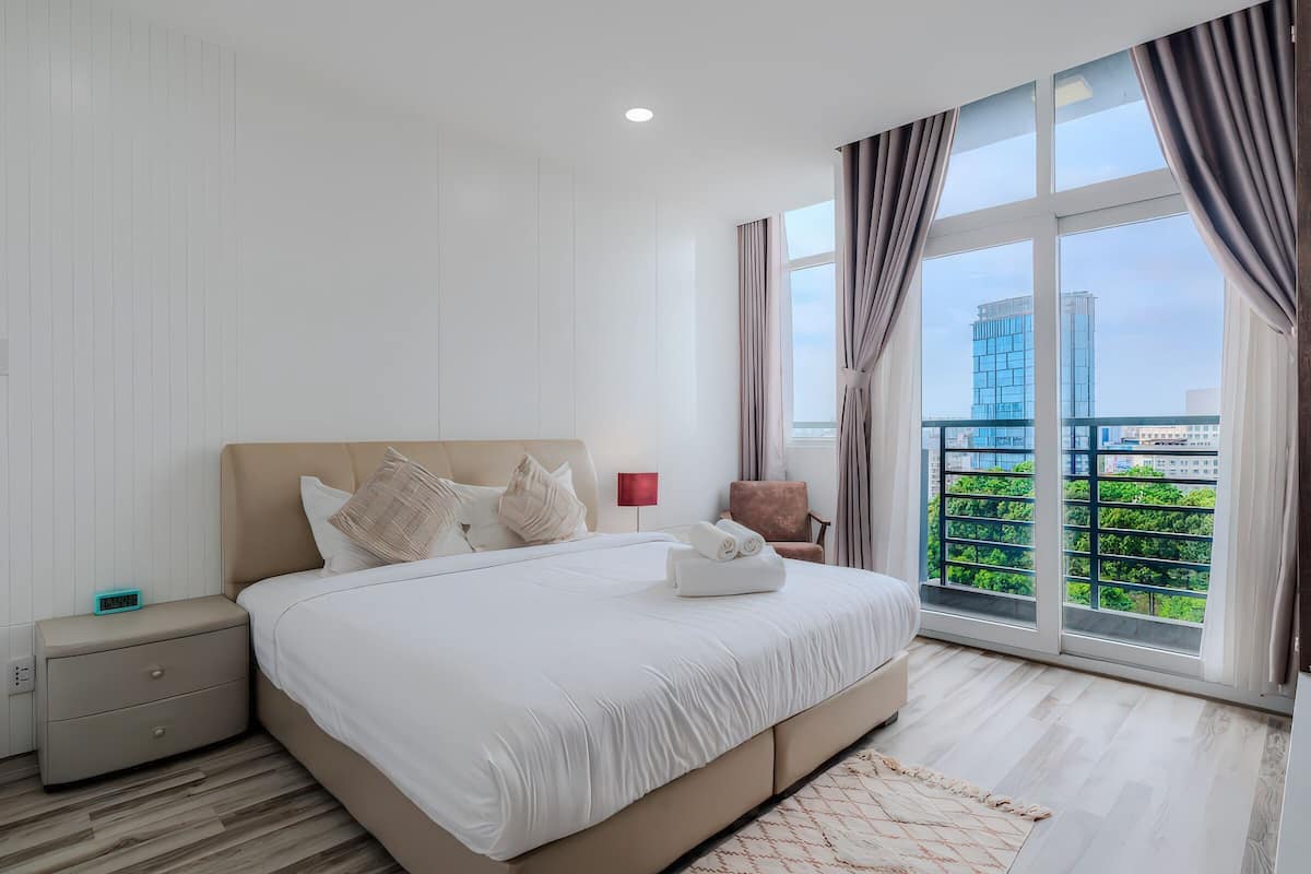 A modern hotel room with a large bed, a nightstand, a red lamp, and a view of a cityscape through large windows.