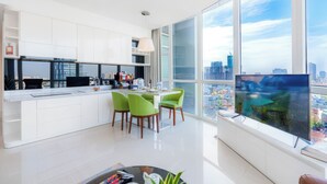 A modern kitchen with white cabinetry, a dining area with green chairs, and a large window with a city view. - Ben Thanh Tower - Luminous Apartments D1 (Ho Chi Minh City)