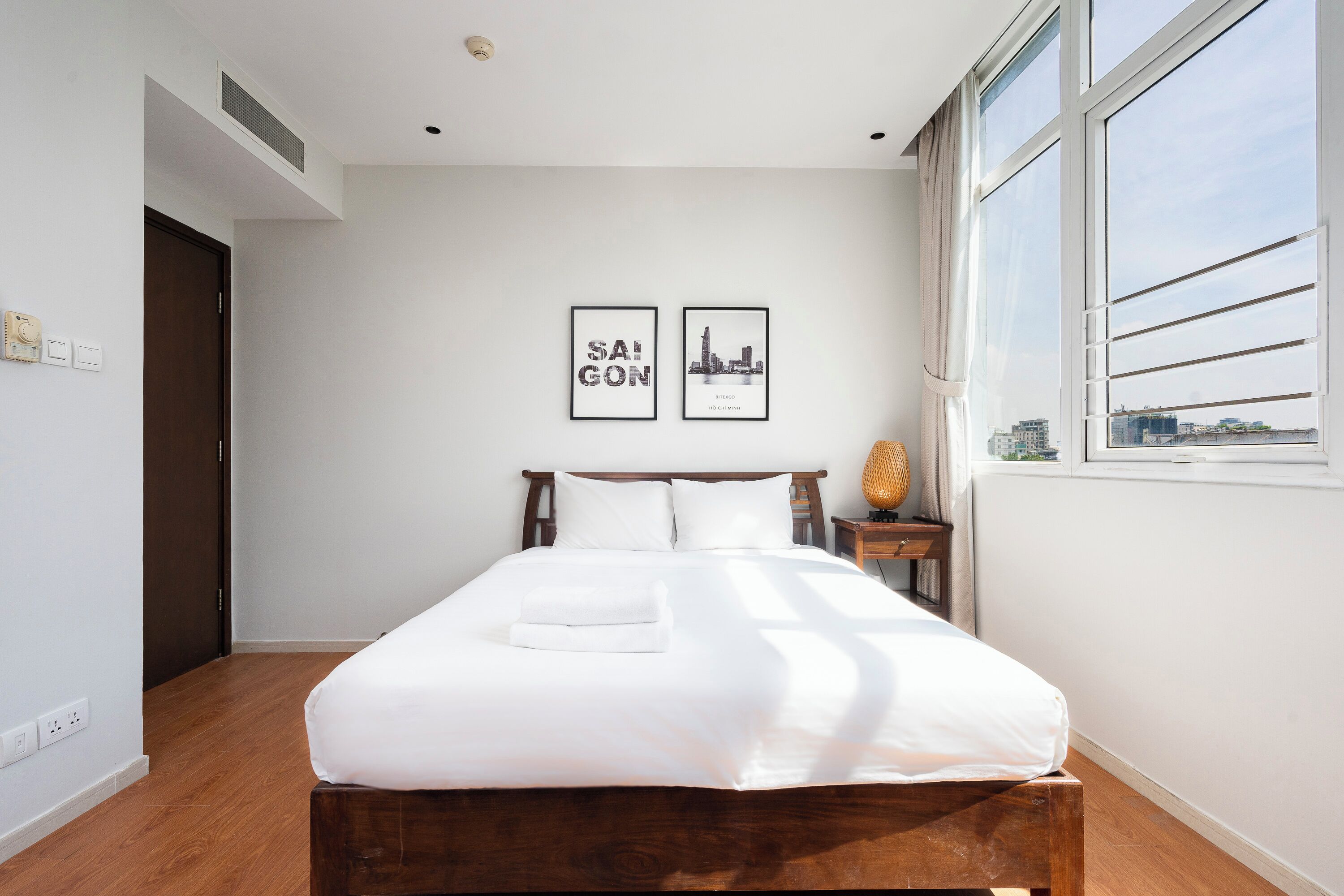 Ein Schlafzimmer mit einem hölzernen Bett, weißer Bettwäsche, einem Stadtbild als Bild und einem Fenster mit Blick auf Gebäude.