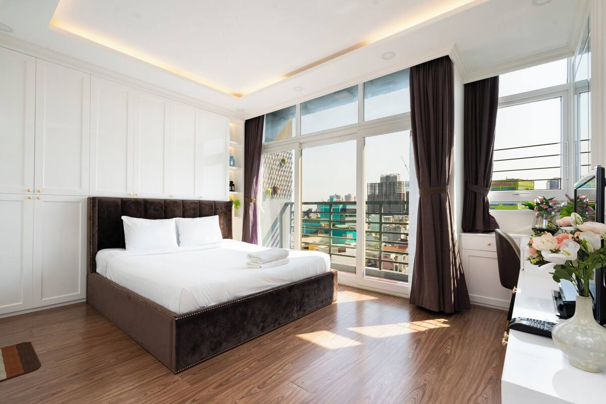 A modern bedroom with a large bed, a desk with a computer, and a view of a cityscape through large windows.