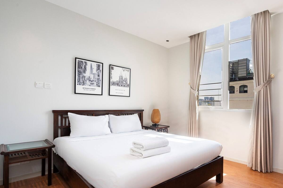 A neatly made bed with white linens, a wooden headboard, and a nightstand with a lamp. Two framed pictures are hung on the wall above the bed.