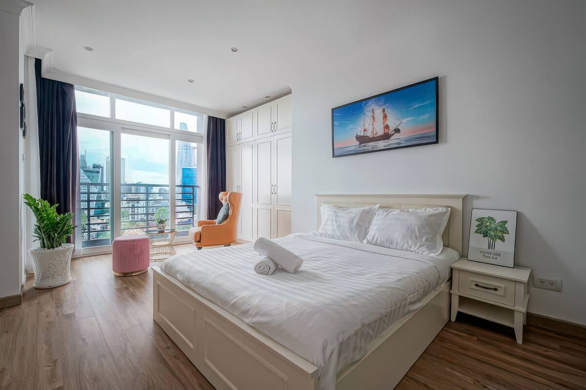 A modern bedroom with a large bed, a city view, a potted plant, and a framed picture on the wall.