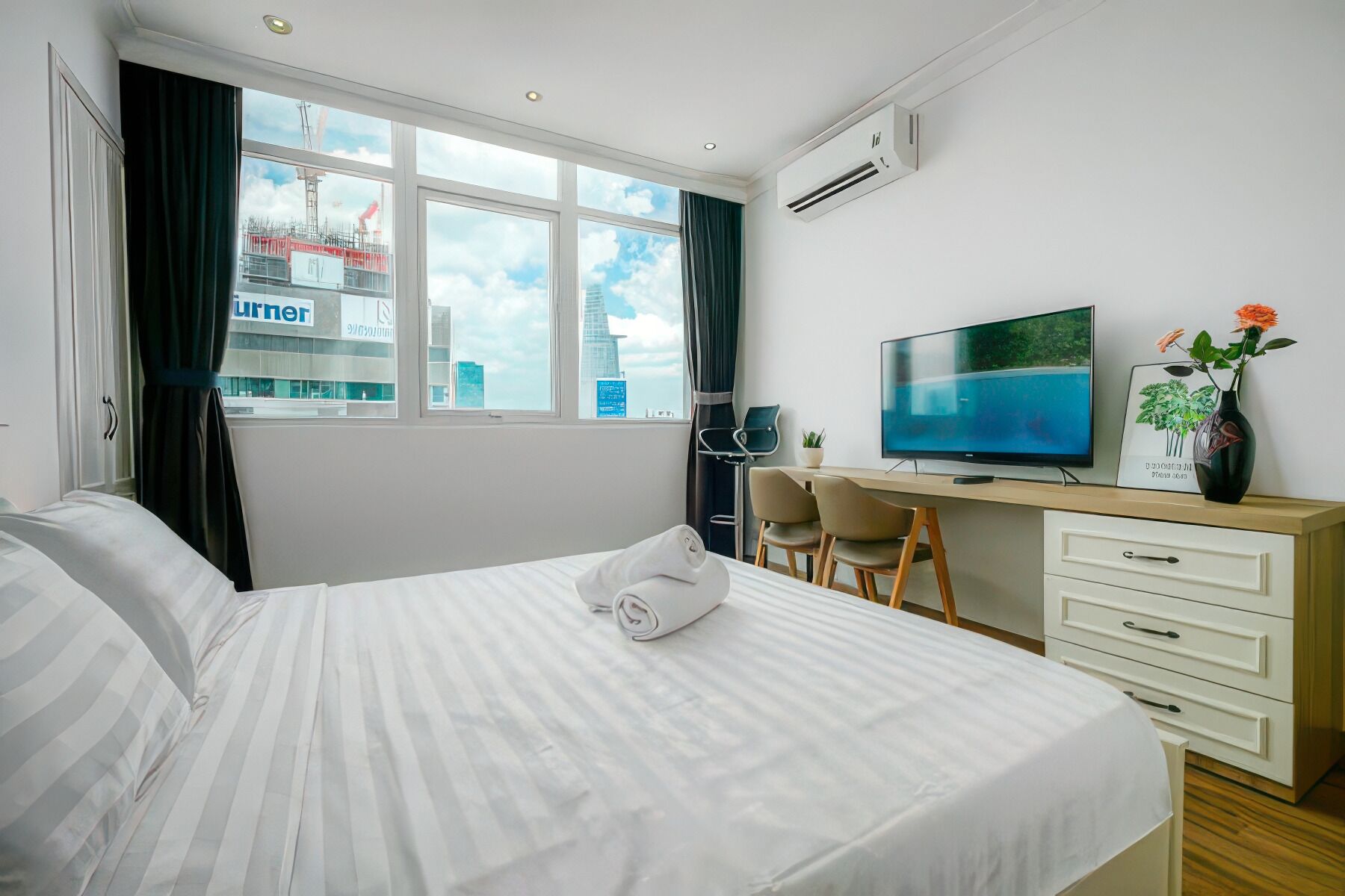 A hotel room with a bed, a desk with a computer, a TV, and a window with a view of buildings.