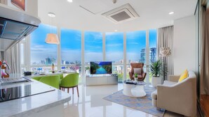 A modern living room with a large TV, green chairs, a white sofa, and a view of the cityscape through large windows. - Ben Thanh Tower - Luminous Apartments D1 (Ho Chi Minh City)
