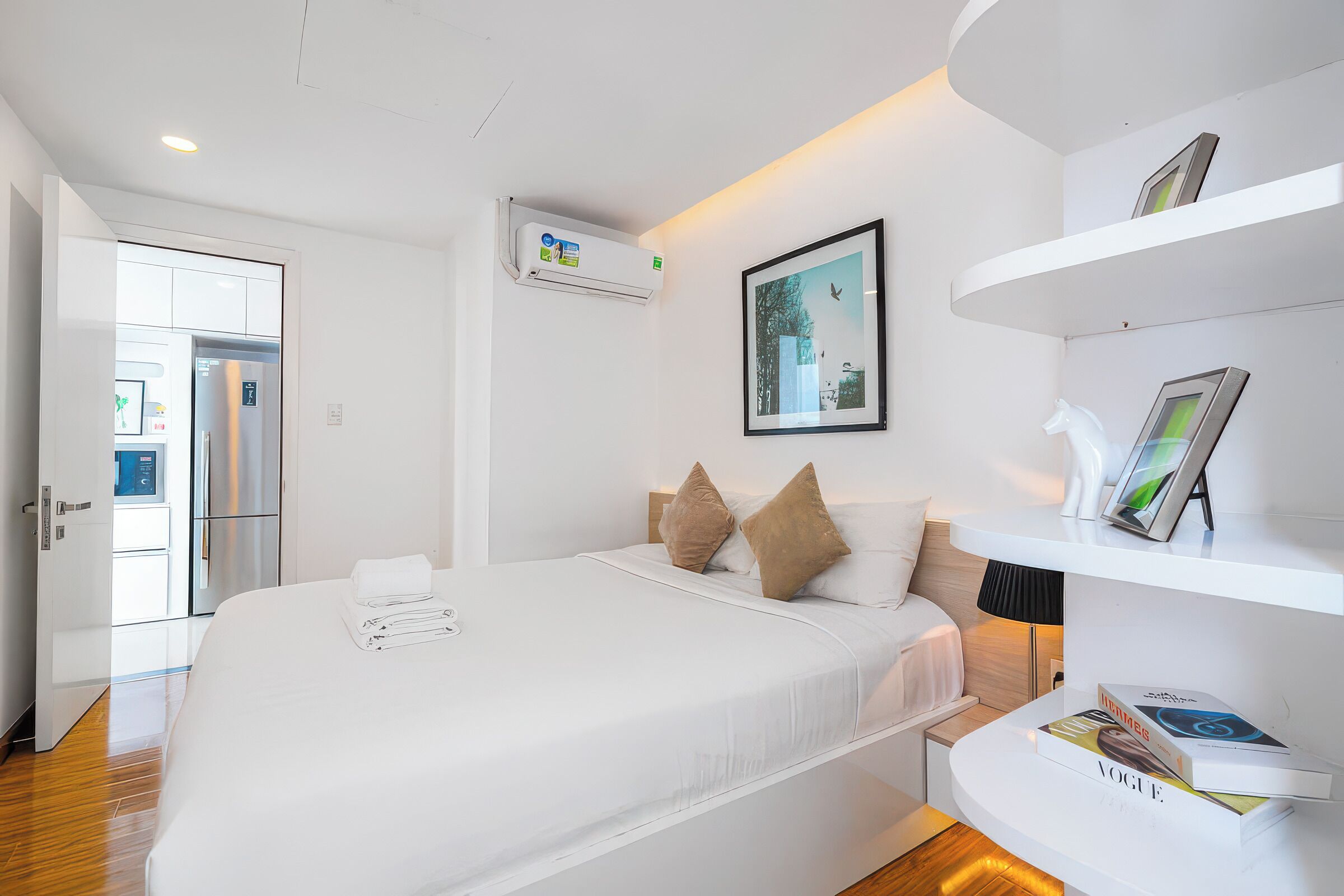 A modern hotel room with a large bed, a bedside table with books, a wall-mounted air conditioner, and a door leading to a kitchen area.