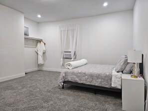 Ein Schlafzimmer mit Bett, Nachttisch mit Lampe, Kleiderschrank und einem Fenster mit Jalousien.
