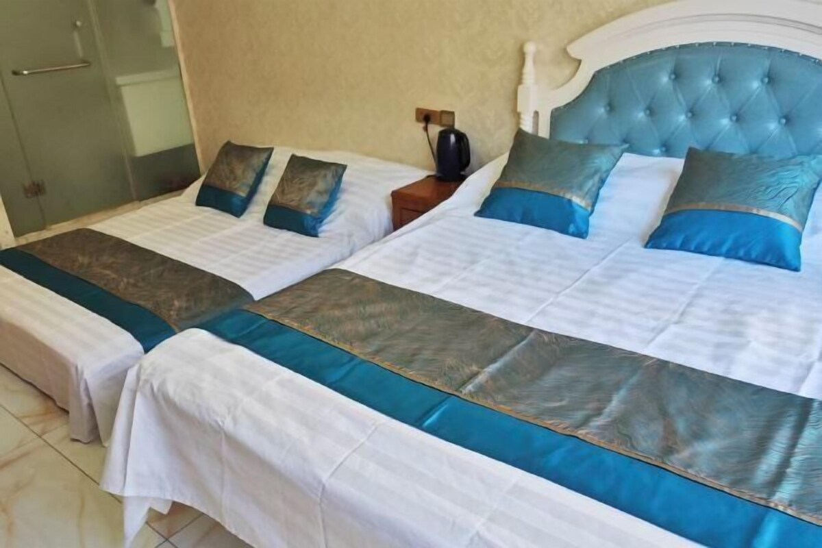 Twee eenpersoonsbedden met wit en teal beddengoed, een houten nachtkastje met een zwart object, en een blauw gestoffeerd hoofdeinde.