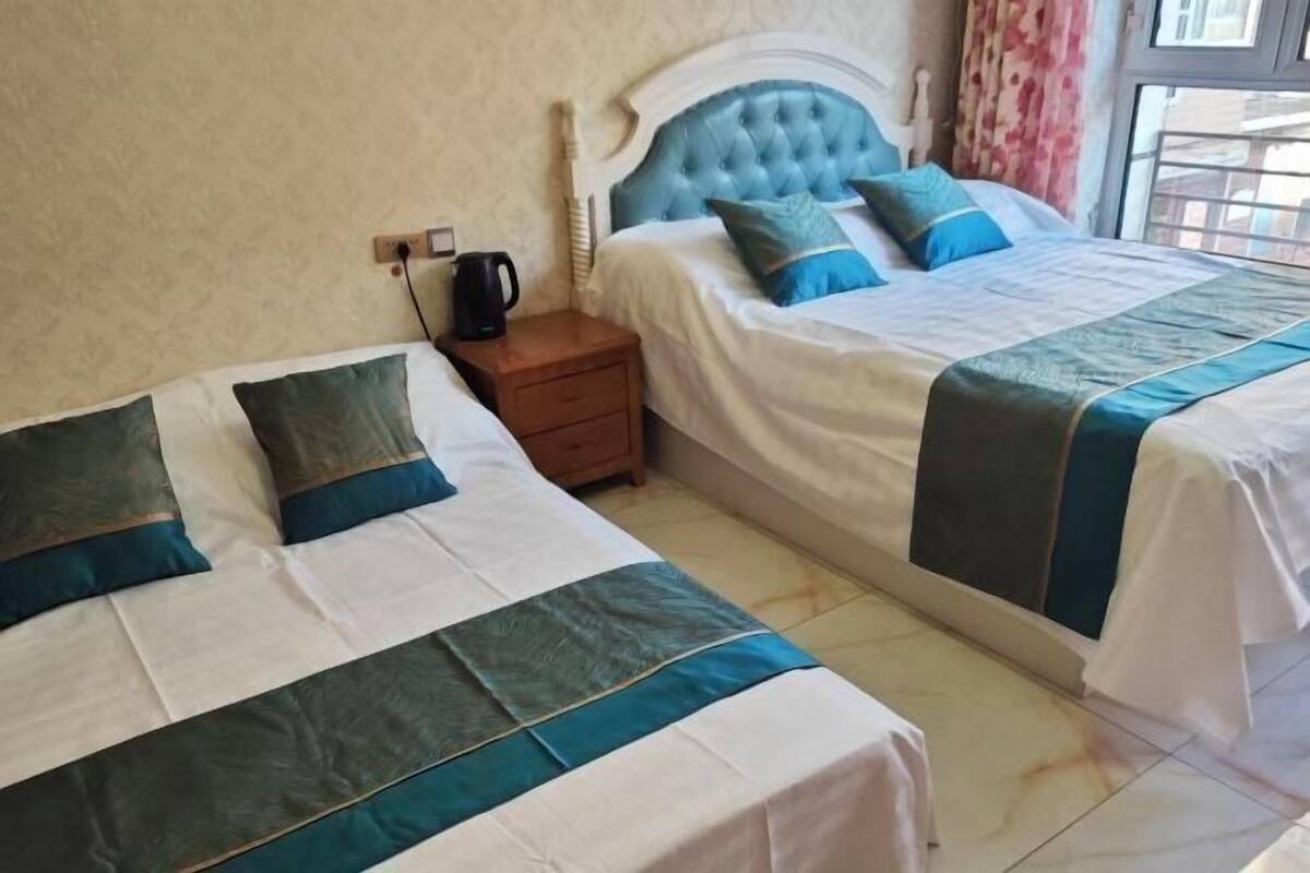 Een hotelkamer met twee bedden, elk met een wit en blauw beddekleed, een houten nachtkastje met een zwart object, en een raam met gordijnen.