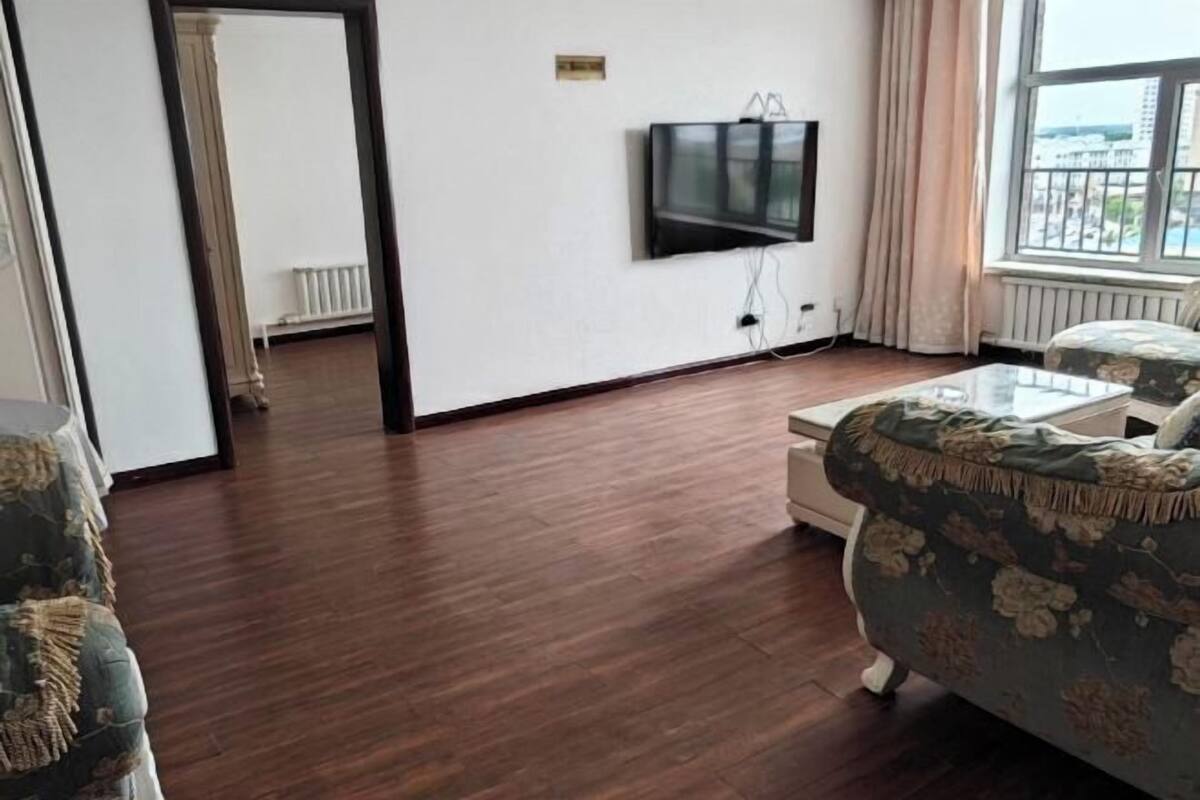 Een kamer met een houten vloer, een bank, een aan de muur bevestigde televisie en een raam met gordijnen.