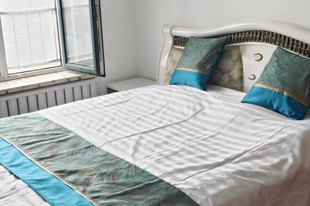 Een netjes opgemaakt bed met een gestreept dekbed en twee kussens, een wit hoofdbord en een raam met uitzicht.