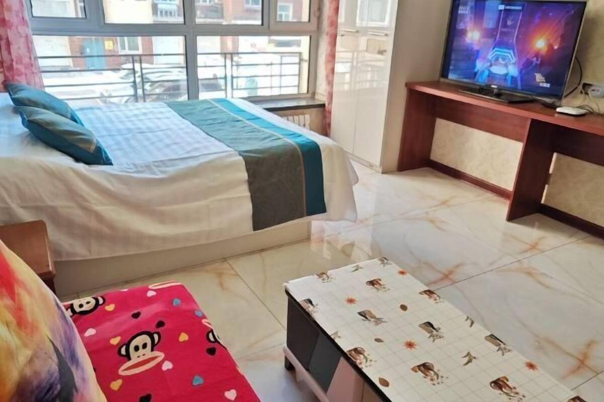 Een slaapkamer met een bed, een televisie op een standaard, een klein tafeltje en een raam met uitzicht op gebouwen.