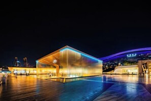 Um edifício moderno com fachada de vidro e área aberta, localizado em frente à orla marítima, com vista para o horizonte da cidade.