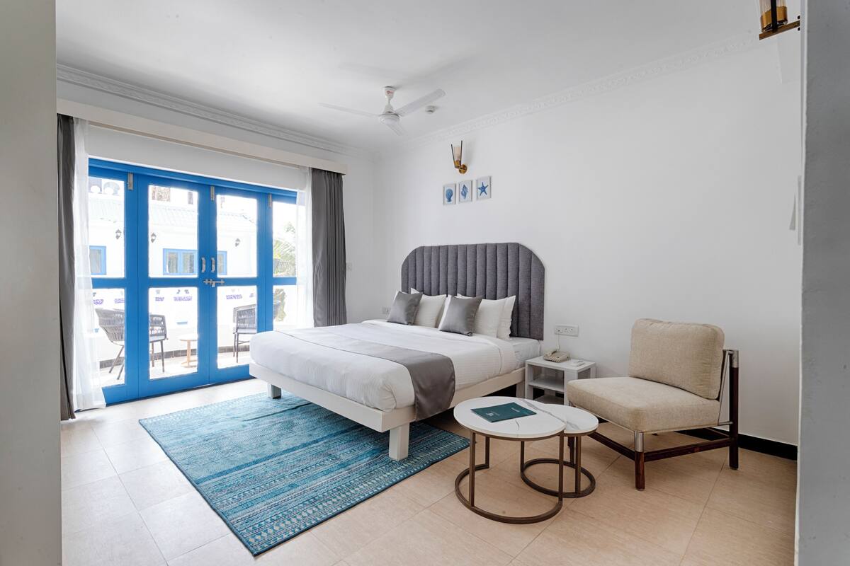 A modern hotel room with a large bed, a bedside table, a chair, and a view through a blue door.