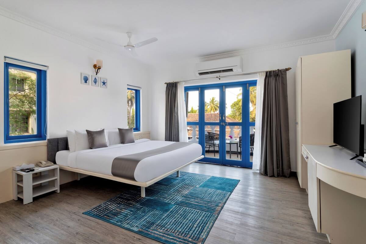 A modern hotel room with a large bed, a desk, a TV, and a balcony with blue doors.