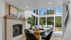 A modern living room with a fireplace, a round wooden table, and a large window overlooking a garden. - Luxury Escape, Hot Tub, Fireplace, Espresso Bar (Ickesburg)