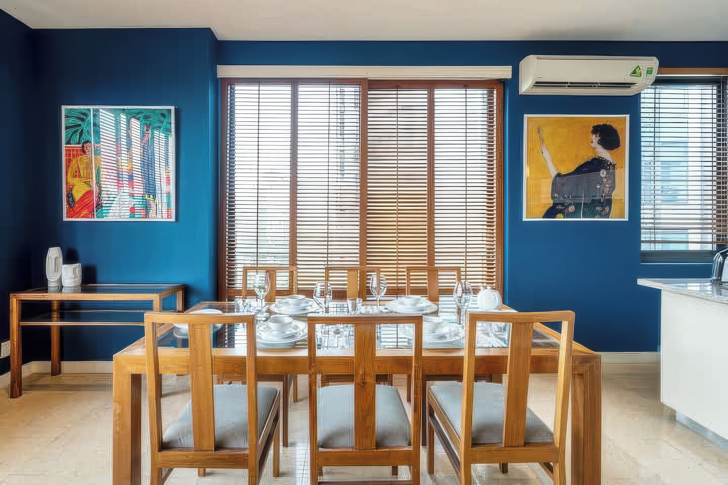 A dining area with a wooden table set for six, flanked by two side tables, in a room with blue walls and large windows with blinds.