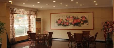 A hotel lobby with a dining area, a large floral painting on the wall, and a large window with curtains.