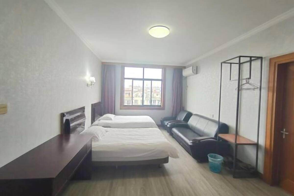 A hotel room with two beds, a sofa, a wooden desk, a window with curtains, a coat rack, and a door.