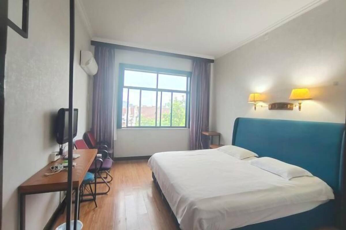 A hotel room with a bed, a desk, a chair, a window with curtains, and a wall-mounted lamp.