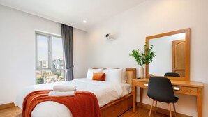 A modern hotel room with a bed, desk, and chair, a large window with a city view, and a mirror on the wall.