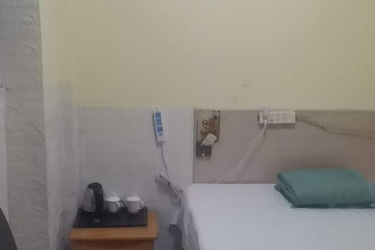 Una cama individual con un almohadón verde, una mesita de noche de madera con tetera y tazas, y un enchufe en la pared.
