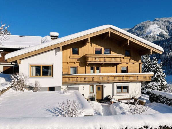 Un chalet en bois à deux étages, doté d’un balcon, entouré d’arbres et de montagnes couverts de neige.